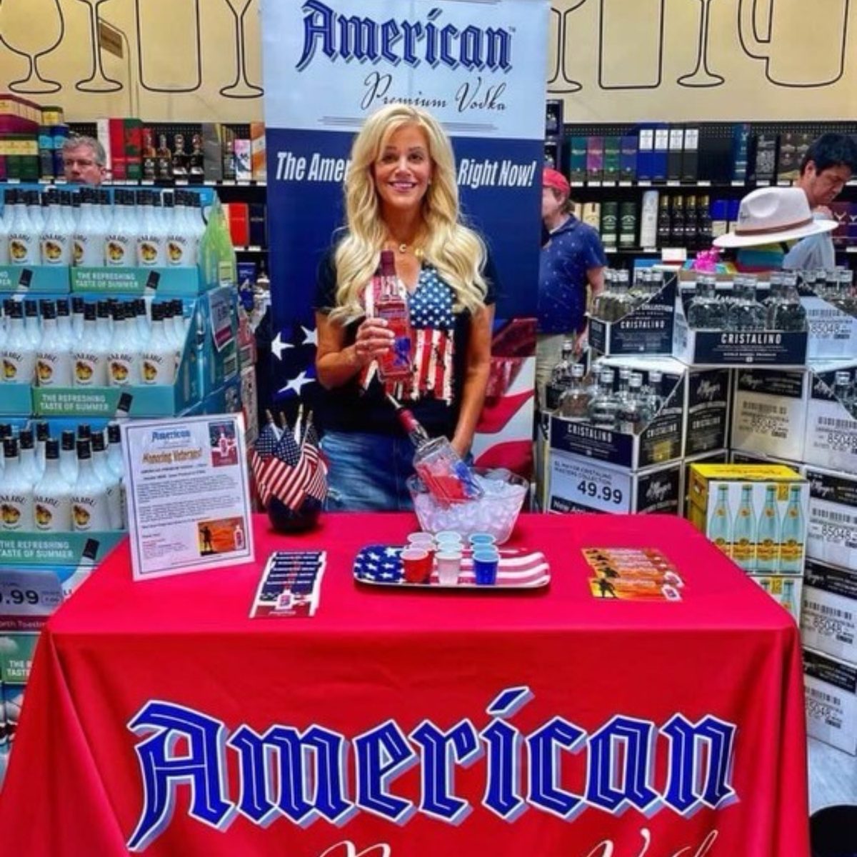 A brand host holding a bottle at a red American Premium Vodka tasting table in a liquor store, with a banner and small flags