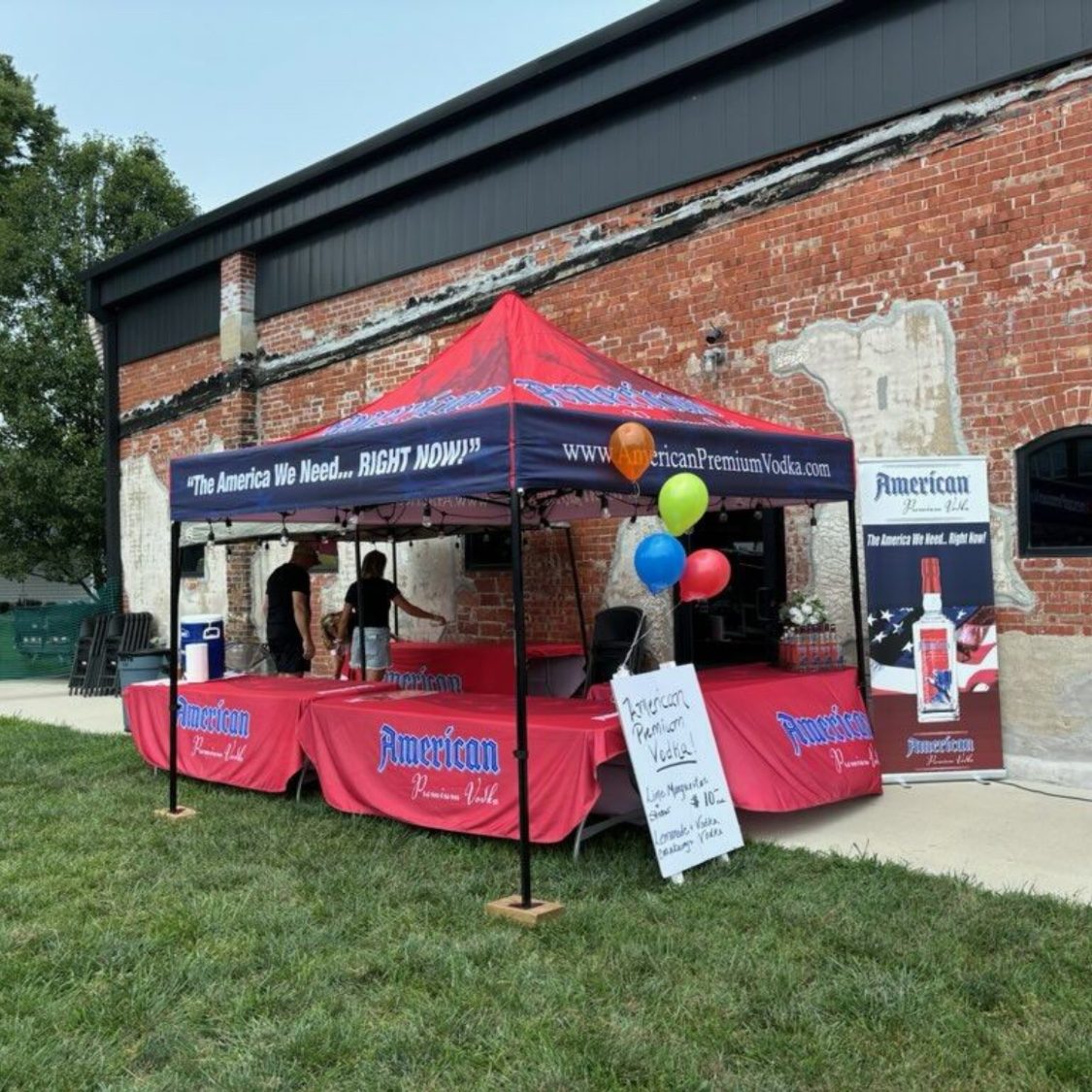 An American Premium Vodka tent with red tables, balloons and a banner outside a brick building
